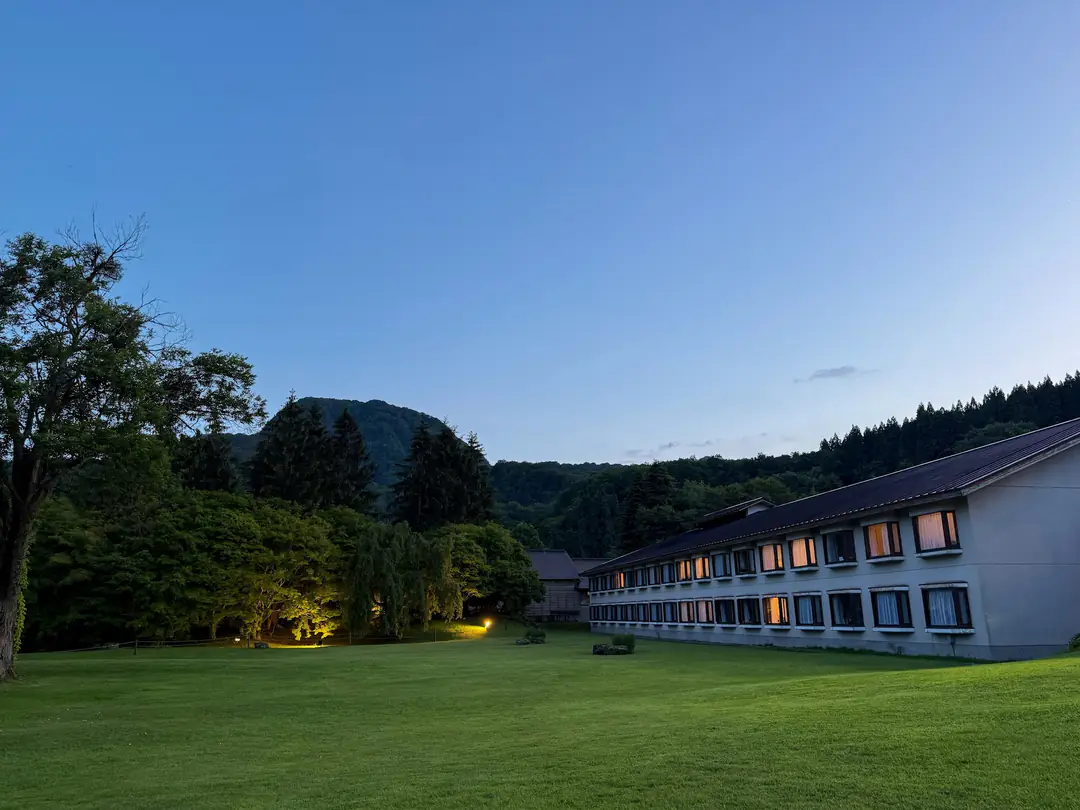 A low building with glowing windows on a lawn, mountains behind at dusk