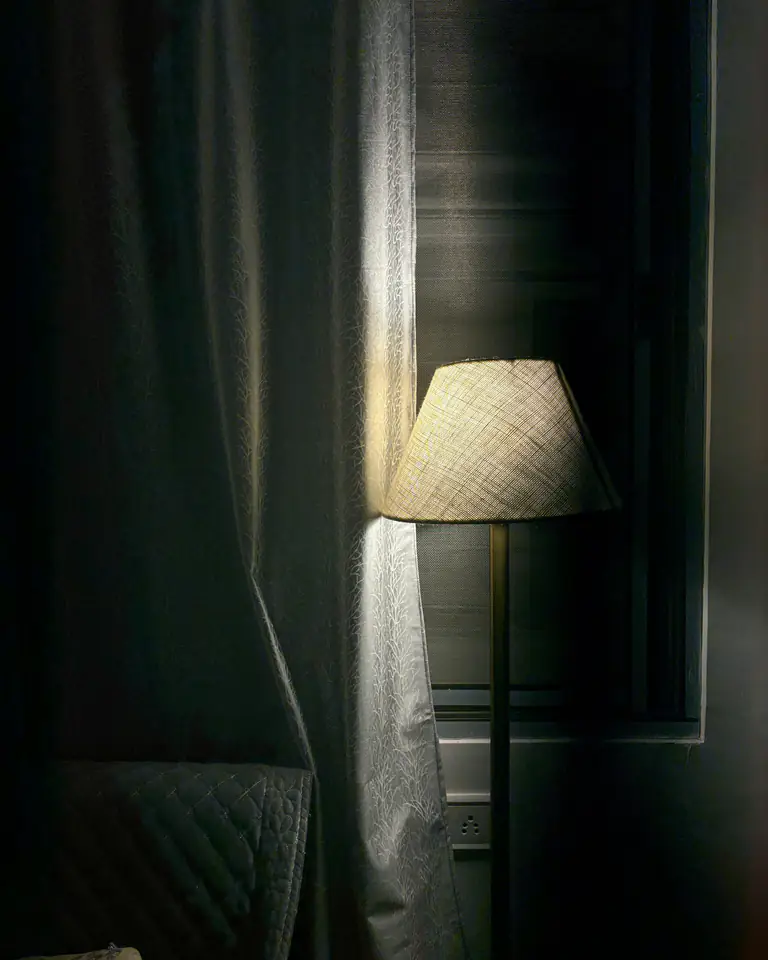 A linen-shaded lamp lit beside a curtained window at night