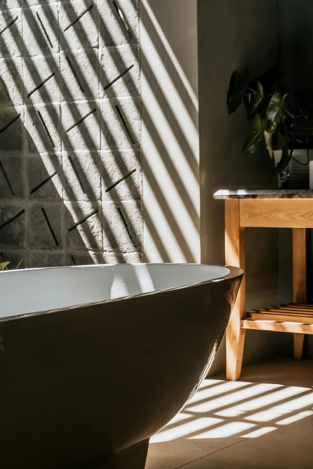 A dark freestanding bathtub beside a stool, striped with sunlight from a window