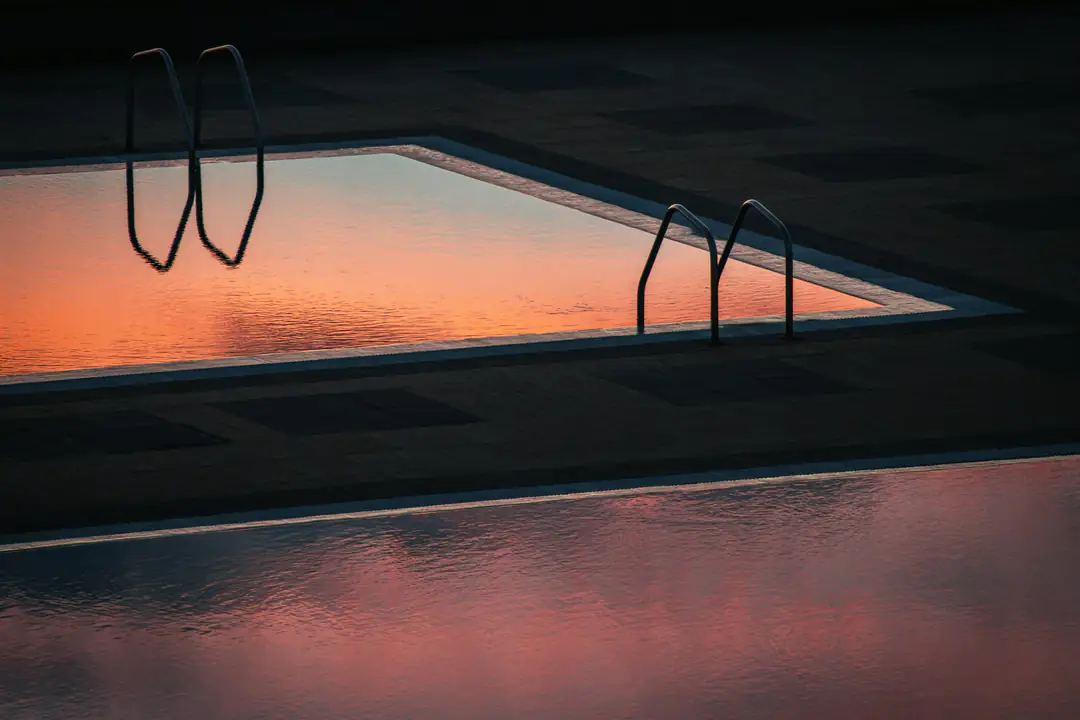 Still pool water reflecting a pink and orange dusk sky, the ladder rails in silhouette